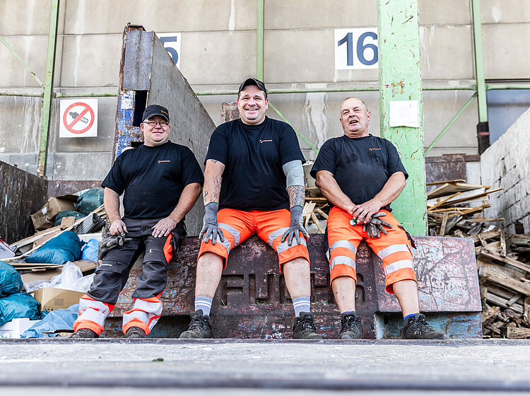 Drei Männer sitzen nebeneinander auf einem Wertstoffhof
